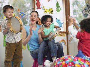 Teacher sits with one student on her lap playing tambourine, other students playing around her.