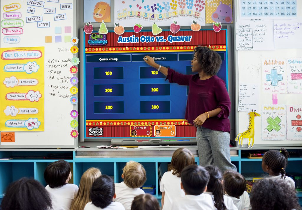 Maestra de preescolar usando Quaver Music en una pizarra interactiva para enseñar a sus alumnos que están sentados frente a ella.