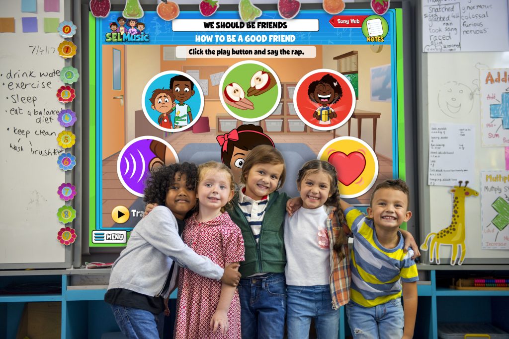 Group of five elementary students stand hugging in front of an interactive whiteboard in a classroom with an activity on friendship displayed.