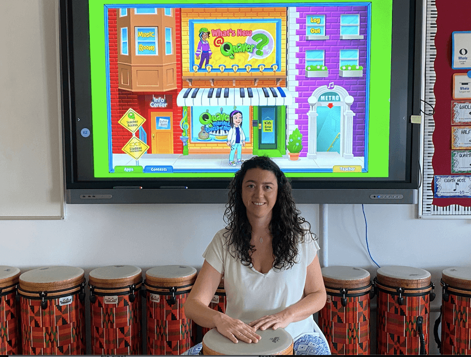 Image of Quaver Music teacher Mari Carmen sitting in her classroom with a drum and Quaver on the screen behind her