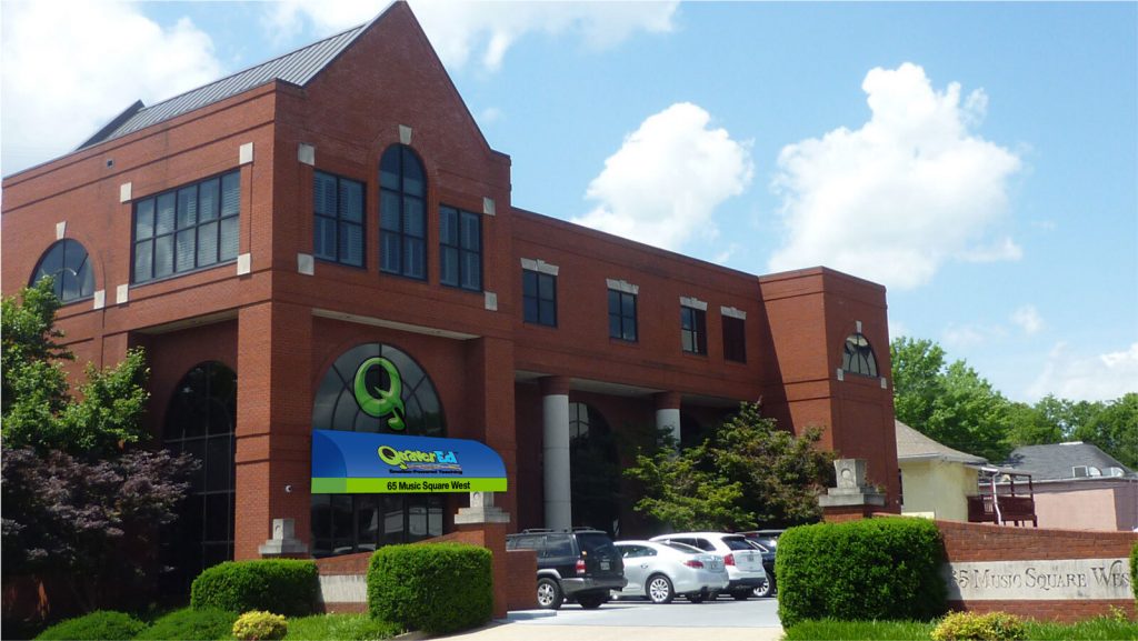 Large brick office building with a QuaverEd logo on a bright blue and green awning. Cars are parked in front of the building on a sunny day. 