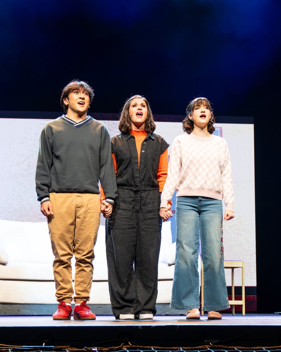 Three cast members stand hand in hand singing on stage
